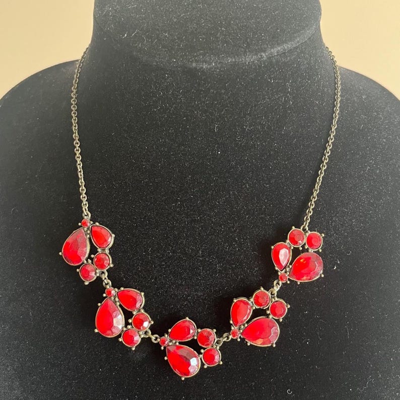 Pu&ograve; includere: Una collana appariscente con grappoli di pietre preziose rosse a forma di goccia e rotonde. Le pietre sono incastonate su una delicata catena color oro antico. La collana &egrave; esposta su uno sfondo nero.