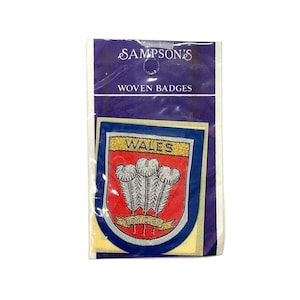 Può includere: Un distintivo tessuto blu e rosso con la parola "WALES" in lettere dorate. Il distintivo presenta tre piume bianche su uno sfondo rosso con un nastro dorato che recita "ICH DIEN". Il distintivo è confezionato in un sacchetto di plastica trasparente con un cartoncino bianco che recita "SAMPSON'S WOVEN BADGES".