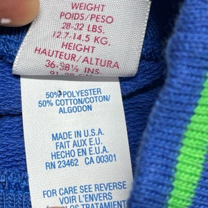 May include: Close-up of a clothing tag with text in English, French, and Spanish. The tag lists weight as 28-32 lbs and 12.7-14.5 kg, and height as 36-38.5 inches. The tag also indicates the garment is 50% polyester and 50% cotton, made in the U.S.A.