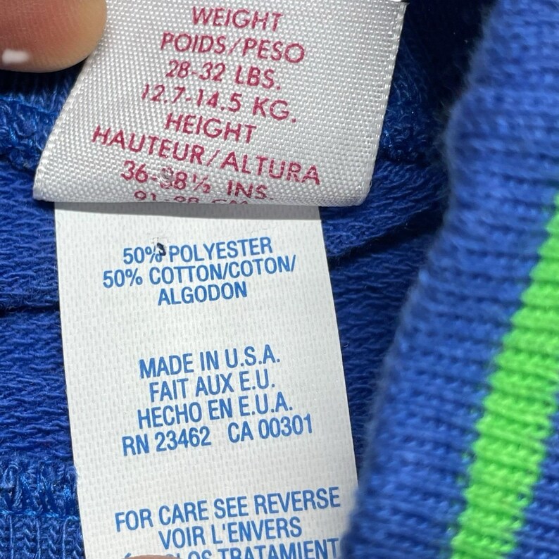 May include: Close-up of a clothing tag with text in English, French, and Spanish. The tag lists weight as 28-32 lbs and 12.7-14.5 kg, and height as 36-38.5 inches. The tag also indicates the garment is 50% polyester and 50% cotton, made in the U.S.A.