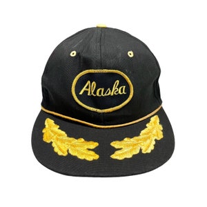 May include: Black baseball cap with a gold embroidered oval patch that says "Alaska" and two gold embroidered oak leaves on the brim.
