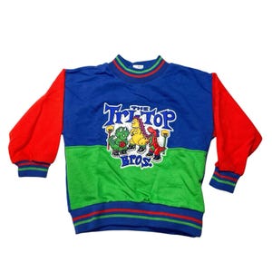 May include: A blue, green, and red sweatshirt featuring a cartoon graphic of three dinosaurs with the text "The Tri-Top Bros." The sleeves are red, and the collar and cuffs have striped detailing. The sweatshirt is likely vintage.