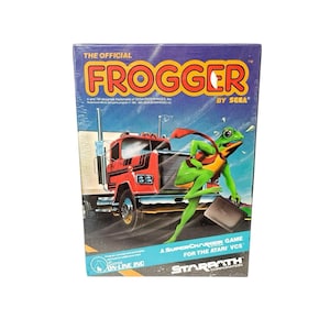 Può includere: Una scatola di videogiochi vintage di Frogger. La scatola presenta un personaggio di rana verde con un mantello rosso e una valigetta, che corre davanti a un camion rosso. Il titolo del gioco è in grandi lettere gialle. La scatola è sigillata in plastica.