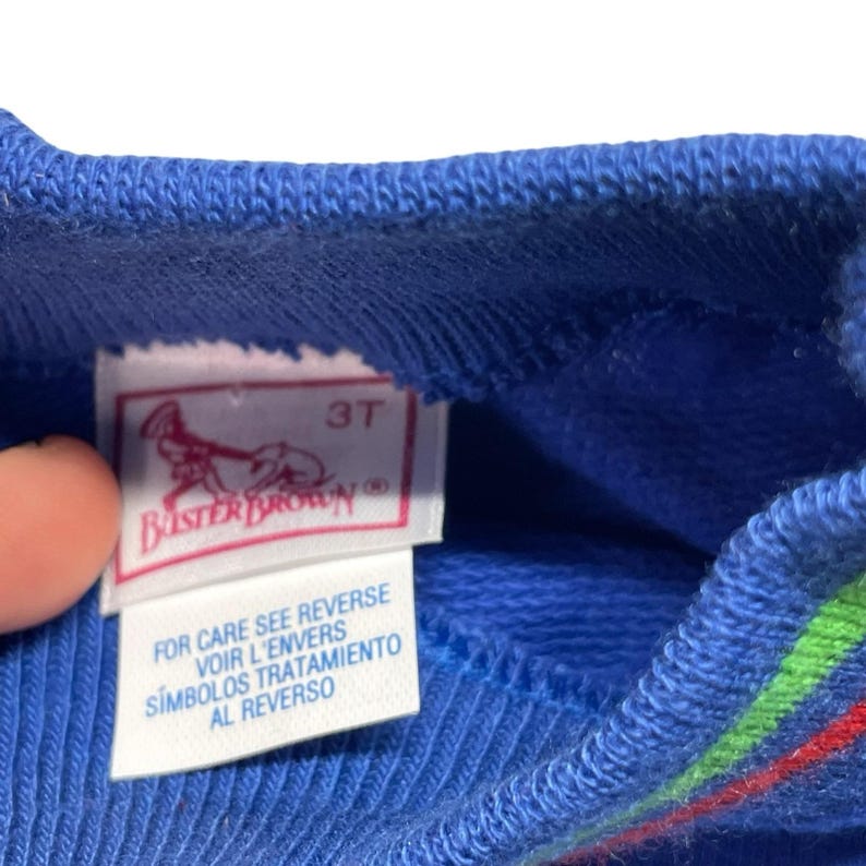 May include: Close-up of a blue children's garment with a tag. The tag is white with red and pink accents, featuring the text "3T" and the Buster Brown logo. The tag also includes care instructions in multiple languages.