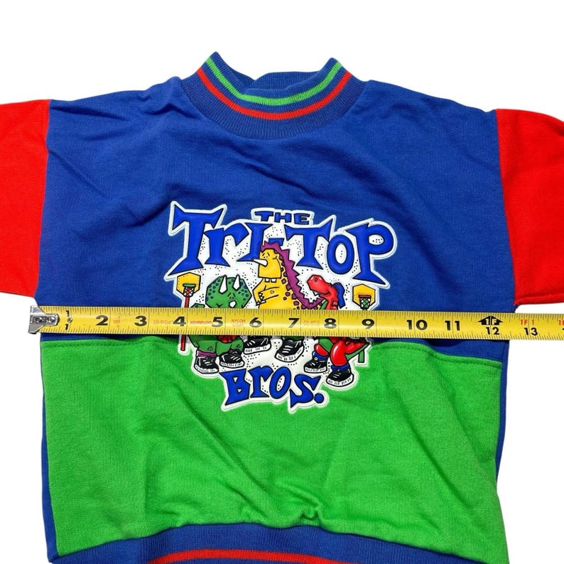 May include: A blue, green, and red sweatshirt with the text "THE Tri-TOP Bros." and cartoon characters. The sweatshirt has a mock neck with red, green, and blue stripes. The sleeve is red, and the body is blue and green.