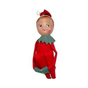 Può includere: Un ornamento di Natale vintage in feltro a forma di elfo con un vestito rosso, un colletto verde e un viso dipinto. L'elfo tiene un rotolo arrotolato nelle sue mani.