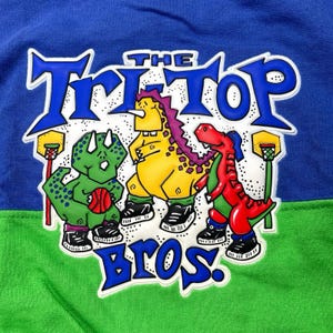 May include: A blue, green, and white graphic tee featuring three cartoon dinosaurs playing basketball. The text "THE TRI-TOP BROS" is displayed in blue. The dinosaurs are green, yellow, and red, each wearing sneakers.