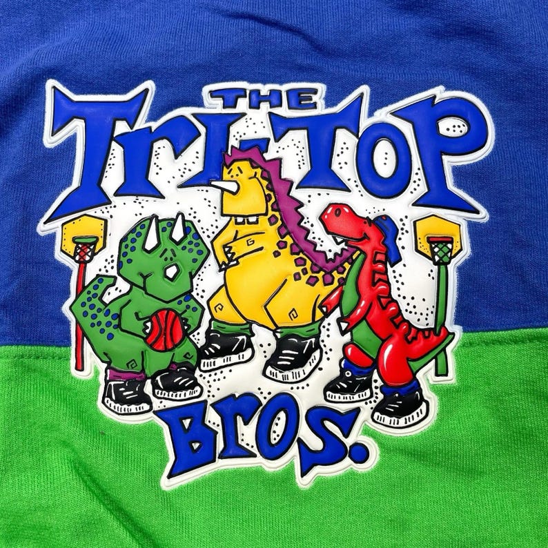 May include: A blue, green, and white graphic tee featuring three cartoon dinosaurs playing basketball. The text "THE TRI-TOP BROS" is displayed in blue. The dinosaurs are green, yellow, and red, each wearing sneakers.