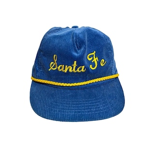 May include: Blue corduroy baseball cap with the embroidered text "Santa Fe" in yellow script. The cap features a yellow rope detail along the brim. A classic, casual accessory.