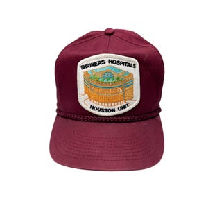 May include: A maroon trucker hat with a white patch featuring the text "Shriners Hospitals Houston Unit" and a graphic of a building.