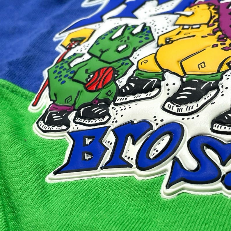 May include: A close-up of a sweatshirt with a color block design in green, blue, and white. The sweatshirt features a graphic of cartoon dinosaurs wearing sneakers and the word "BROS" in blue and white.