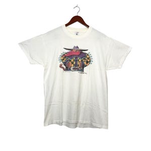 May include: A white t-shirt with a cartoon illustration of a cowboy wearing a purple shirt, a red bandana, and a black hat. The cowboy is holding a lasso and has a cat sitting on his shoulder. The text "The Cat That Got Away" is printed below the illustration.