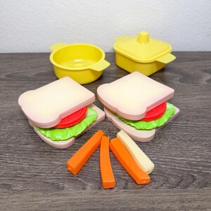 May include: A set of plastic play food including two sandwiches with bread, lettuce, and tomato slices, two yellow containers with one lid, and four rectangular sticks in orange and off-white colors. The items are arranged on a flat surface.