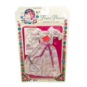 May include: A Flower Princess doll fashion set for 29.2 cm dolls. The set includes a pink and white striped dress with a pink ribbon, a matching purse, and a collector sticker. The packaging features floral designs and the text "Flower Princess".