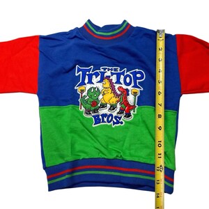 May include: A vintage children's sweatshirt with a blue, green, and red color block design. The sweatshirt features the text "The Tri-Top Bros" with cartoon dinosaur characters. The length of the sweatshirt is approximately 14 inches.