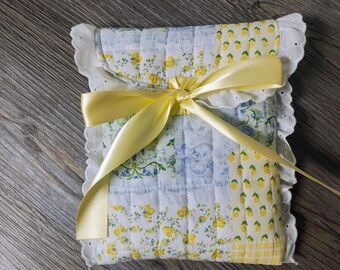 May include: A small quilted pouch with a yellow floral pattern and a yellow satin ribbon tied around it. The pouch has a white lace trim.