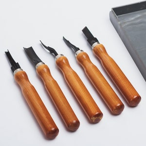 Puede incluir: Conjunto de cinco herramientas de talla de madera con mangos de madera y cuchillas de metal negro. Las herramientas están en una caja de plástico gris con el texto "НАБОР РЕЗЦОВ" impreso en la caja.