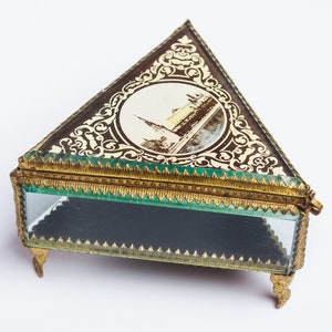 May include: A triangular glass trinket box with gold trim and a decorative image of a building on the lid. The box has three feet and a hinged lid.