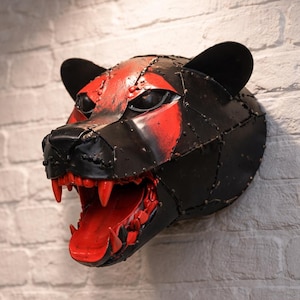 Könnte beinhalten: Eine Wandskulptur eines Pantherkopfes, gefertigt aus vernieteten schwarzen Metallplatten. Der Panther weist leuchtend rote Akzente um die Augen auf und ein weit geöffnetes Maul, das rote Zähne und eine rote Zunge zeigt, was einen wilden Ausdruck erzeugt.