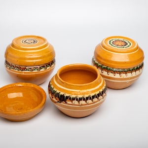 Op de afbeelding: Een verzameling handgemaakte keramische potten. De stukken zijn voornamelijk oranje met decoratieve banden in wit, groen en bruin. De set bevat potten met deksel en een kleine kom, allemaal met een rustieke, ambachtelijke uitstraling.
