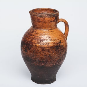 May include: A brown earthenware pitcher with a wide mouth and a single handle. The pitcher has a rustic, aged appearance with a dark brown glaze.