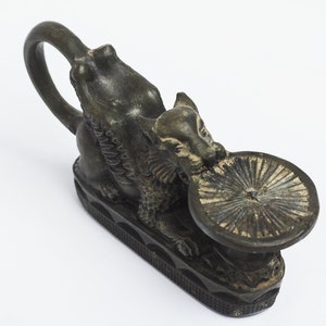 May include: A black metal candle holder shaped like a mythical creature with a round, flat top. The creature has wings and a tail, and its head is turned to the side. The candle holder is sitting on a white surface.