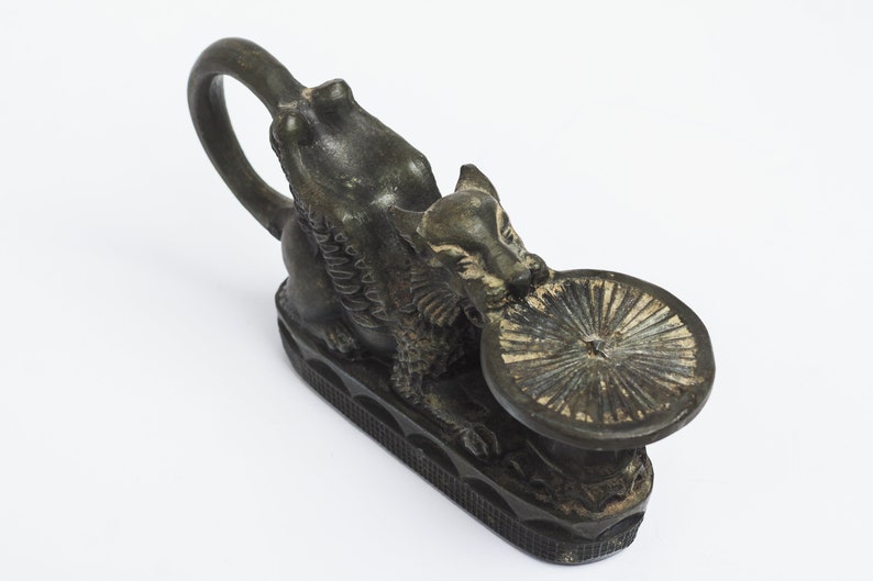 May include: A black metal candle holder shaped like a mythical creature with a round, flat top. The creature has wings and a tail, and its head is turned to the side. The candle holder is sitting on a white surface.