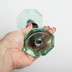 Może przedstawiać: Przezroczysty, jasnozielony szklany korek do butelki o ośmiokątnym kształcie. Korek ma srebrną śrubę pośrodku i ciemnoszary trzpień. Szkło ma lekki odcień i jest trzymane w dłoni.