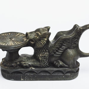 May include: A bronze winged griffin sculpture with a circular dish on its back. The griffin is holding the dish in its mouth. The sculpture has a detailed design and a dark patina.