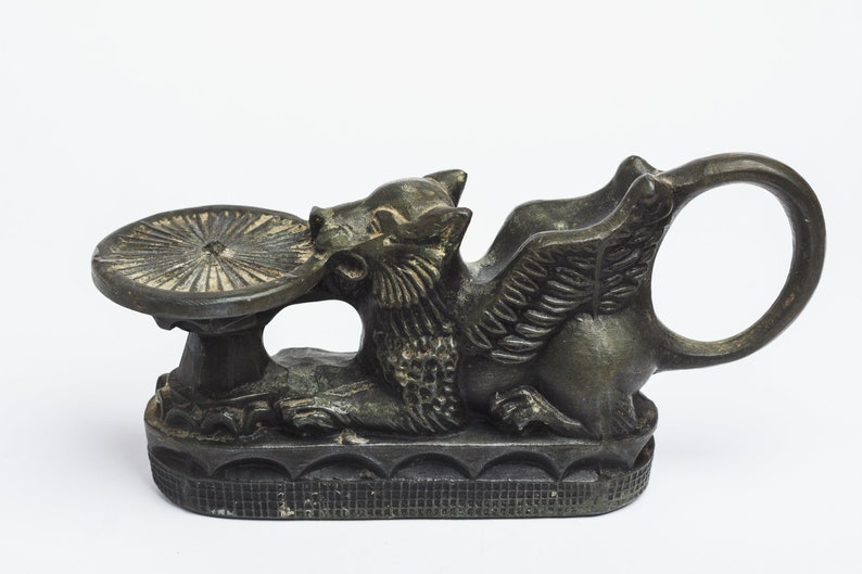 May include: A bronze winged griffin sculpture with a circular dish on its back. The griffin is holding the dish in its mouth. The sculpture has a detailed design and a dark patina.