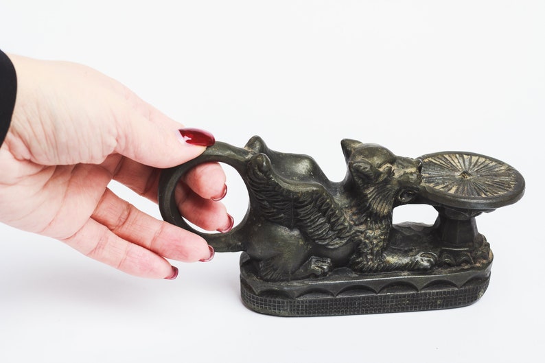 May include: A bronze winged griffin sculpture with a round, flat top. The griffin is holding the top in its mouth and has a ring on its back.