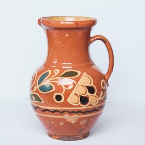 Op de afbeelding: Een bruine keramische kan met een bloemenmotief in tinten blauw, groen, geel en zwart. De kan heeft een brede basis en een smalle hals met een handvat.