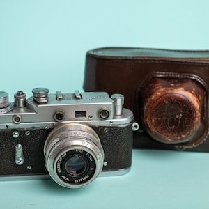 Pode incluir: Uma câmera de filme vintage de 35 mm prata e preta com uma capa de couro. A câmera tem uma lente etiquetada como "Industar-50".