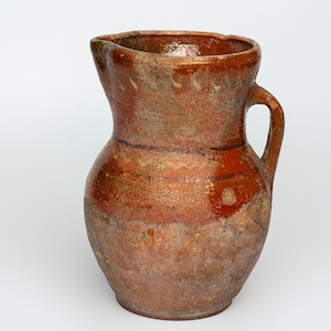 May include: A brown earthenware pitcher with a wide mouth and a single handle. The pitcher has a rustic, handmade appearance and is glazed in a reddish-brown color.