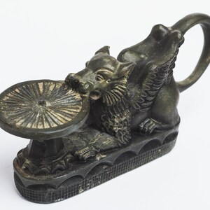 May include: A black metal incense burner shaped like a mythical creature with wings and a round dish on top. The creature is holding the dish in its mouth.