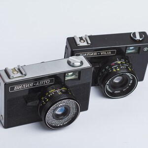 May include: Two black film cameras with silver accents. The camera on the left has the text "ВИЛИЯ-АВТО" on the front. The camera on the right has the text "ВИЛИЯ-VIIIA" on the front.