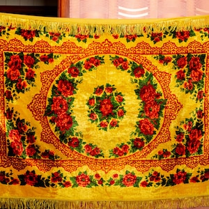 May include: A yellow velvet textile with a floral design. The textile features a central floral medallion surrounded by red roses and green leaves. The border is a darker shade of yellow with intricate patterns, and the edges have a fringe.
