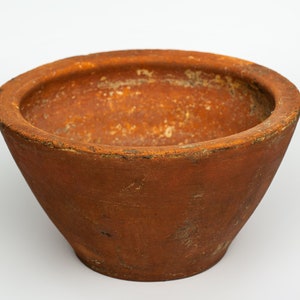 May include: A terracotta bowl with a wide, rounded base and a slightly flared rim. The bowl is a deep reddish-brown color and has a rough, textured surface.