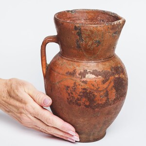 Puede incluir: Una jarra de barro marrón con asa. La jarra tiene un aspecto desgastado y rústico con un esmalte marrón y naranja moteado.
