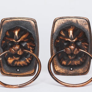 以下が含まれることがあります： ライオンの頭のデザインが施されたアンティーク調のタオルリングが2つ。リングは金属製で、ダークブラウンの仕上げです。