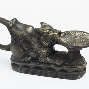 May include: A bronze sculpture of a winged griffin holding a circular dish. The griffin has a detailed, textured surface and a ring handle. The sculpture is on a white background.