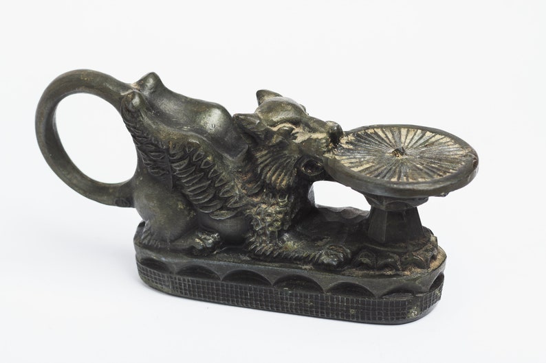 May include: A bronze sculpture of a winged griffin holding a circular dish. The griffin has a detailed, textured surface and a ring handle. The sculpture is on a white background.