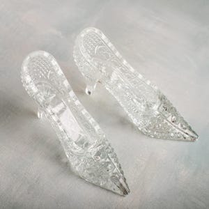 Puede incluir: Dos zapatos de tacón alto de cristal transparente con intrincados patrones tallados. Los zapatos tienen puntas afiladas y tacones pequeños. El cristal tiene una calidad brillante y reflectante, lo que sugiere un objeto decorativo.