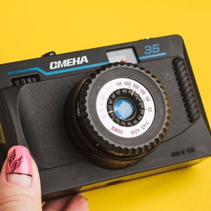 Puede incluir: Cámara de película Smena 35 negra con lente circular y correa negra. La cámara tiene la palabra "Smena" en azul y el número "35" en la parte superior. La lente tiene números y marcas blancas. Sobre fondo amarillo.
