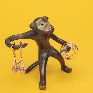 Puede incluir: Una figurita de mono de bronce que sostiene un par de pendientes colgantes con piedras rosas y un juego de tres anillos de oro.