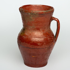 May include: A red earthenware pitcher with a handle. The pitcher has a slightly chipped rim and is glazed with a reddish-brown glaze.