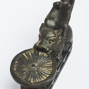 May include: A dark bronze incense burner shaped like a mythical creature with a round, decorative top. The creature is holding its head up and has a ring on its back.