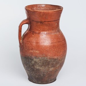 May include: A terracotta pitcher with a wide mouth and a single handle. The pitcher has a reddish-brown glaze and a darker brown base.