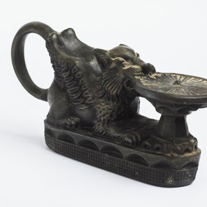 May include: A black bronze sculpture of a winged lion holding a circular plate. The lion is sitting on a base with a decorative pattern.