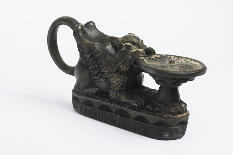 May include: A black bronze sculpture of a winged lion holding a circular plate. The lion is sitting on a base with a decorative pattern.